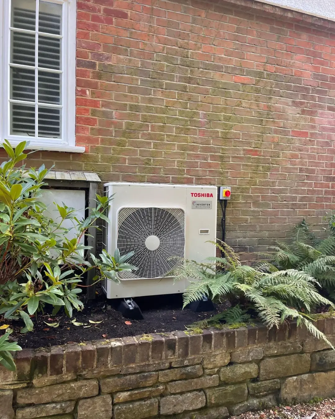 Outdoor unit sited discreetly in a planted border beside a brick house